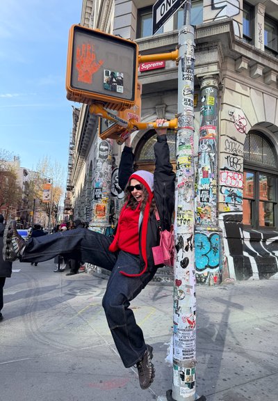 Persona con sudadera roja con capucha y chaqueta negra cuelga de un poste de señal peatonal cubierto de pegatinas en una esquina de calle de la ciudad cubierta de graffiti.