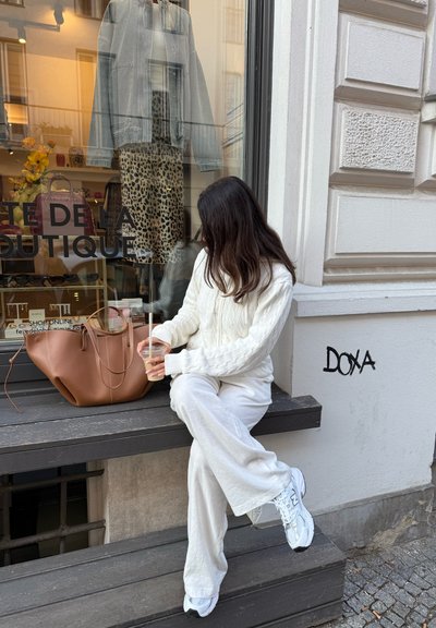 Una persona con unsuéter de punto blanco y pantalones de chándal claros se sienta en un borde de madera, sosteniendo una bebida. A su lado descansa un bolso de cuero marrón.