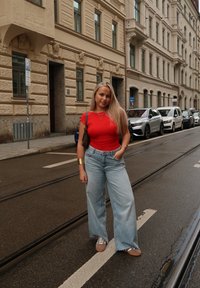 Rode aansluitende top, lichtblauwe wijde jeans, bruine sneakers. Lang blond haar; staat op een straat met geparkeerde auto's en een gebouw op de achtergrond.