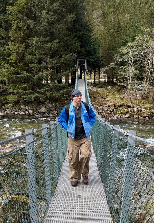 Personne portant une veste bleue et un pantalon cargo marchant sur un pont suspendu métallique étroit au-dessus d'une rivière, entourée d'une forêt dense.