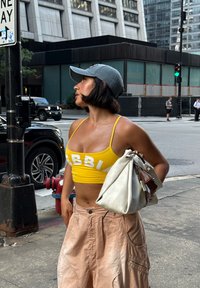 Gul crop top med hvit tekst, ferskenfarge vide bukser og en hvit veske. Utendørs urban setting med en bybakgrunn.