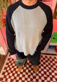 Personne portant un pull côtelé noir et blanc, un pantalon foncé et des bottes beige, tenant une casquette de baseball camouflage sur un tapis à carreaux rouge et blanc.