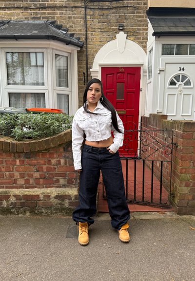 Chaqueta de denim blanca corta, pantalones vaqueros holgados oscuros y botas de trabajo amarillas. De pie cerca de una puerta principal roja con una pared de ladrillo y vegetación.
