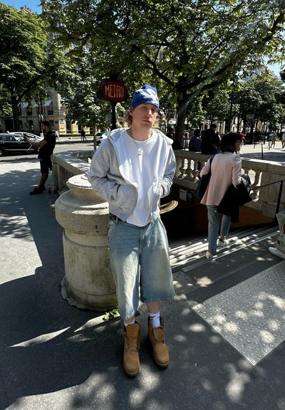 Sudadera gris claro sobre una camiseta blanca, gorro azul, pantalones cortos de mezclilla oversize, calcetines blancos, botas de trabajo marrones. Entorno urbano con vegetación.