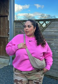 Felpa rosa con logo Nike, borsa a tracolla beige e pantaloni mimetici. Lo sfondo presenta una recinzione in legno e cielo azzurro.