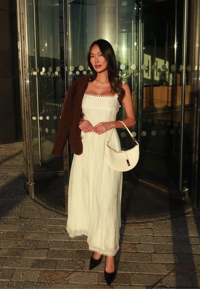 Mujer con un vestido sin mangas color crema, sosteniendo un bolso beige y una chaqueta marrón, de pie frente a unas puertas giratorias de vidrio, bajo la luz del sol.