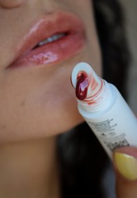 Lucidalabbra in un tubo bianco, con una tonalità rosa lucida sulle labbra e un'intensa tonalità rossa sulla punta. Superficie testa visibile sul tubo.