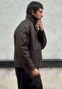Brown leather jacket with a high collar, featuring a smooth texture and structured design. Worn by a person, side profile shown.