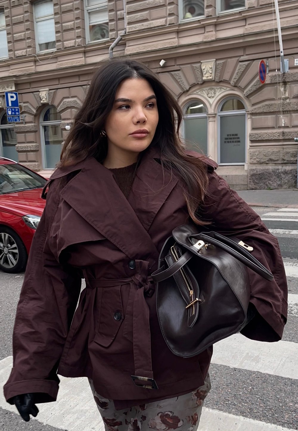 Giacca esterna color marrone scuro con vita con cintura, ampi revers e tasche laterali. Borsa a mano in pelle scura con dettagli dorati. Ambientazione urbana all'aperto.