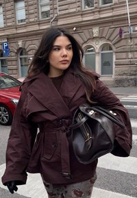 Giacca esterna color marrone scuro con vita con cintura, ampi revers e tasche laterali. Borsa a mano in pelle scura con dettagli dorati. Ambientazione urbana all'aperto.