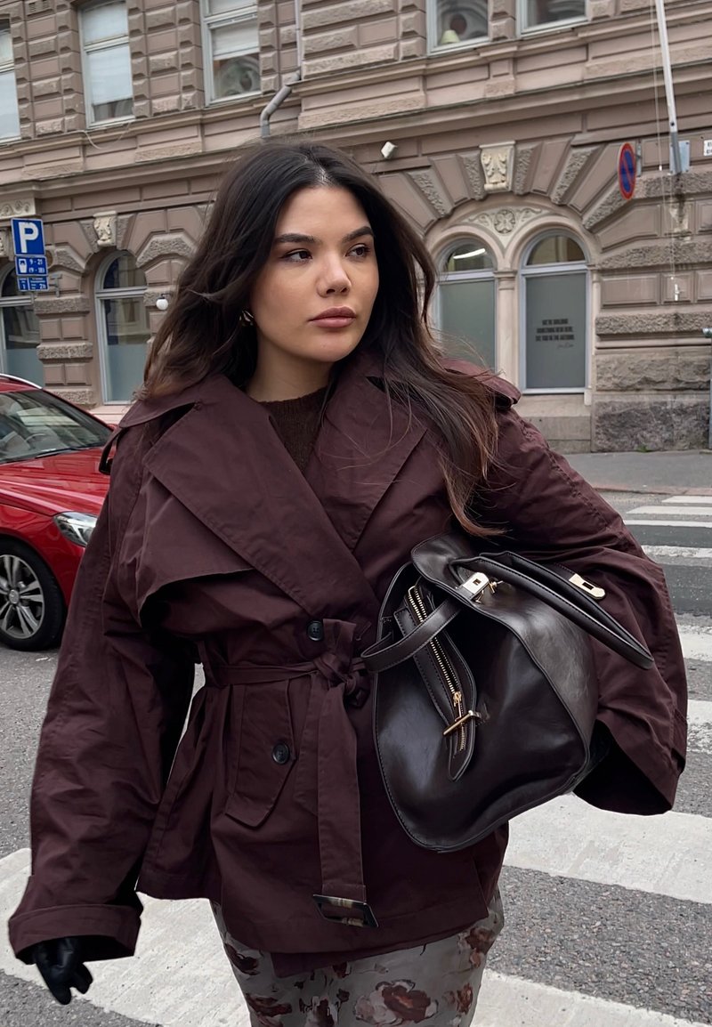 Giacca esterna color marrone scuro con vita con cintura, ampi revers e tasche laterali. Borsa a mano in pelle scura con dettagli dorati. Ambientazione urbana all'aperto.
