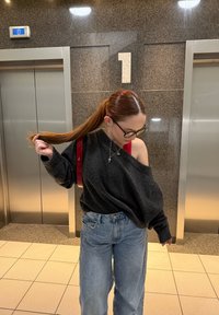 Zwarte oversized trui, lichtblauwe spijkerbroek en een rode tas. Lang bruin haar vastgebonden. Lift met nummer 1 in een moderne omgeving.