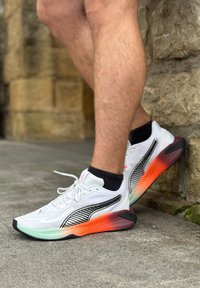 Biele športové topánky s gradientovou podrážkou prechádzajúcou z čiernej do oranžovej farby, s čiernymi pruhmi a textúrovaným vrchom. Čierne členkové ponožky.