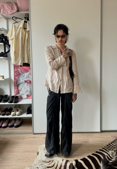 Camisa de lino beige claro con mangas acampanadas, combinada con pantalones negros de corte holgado. Los accesorios incluyen gafas de sol negras y un bolso de hombro.