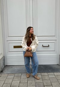 Cremefarvet, tekstureret jakke over en brun top, sammen med løse blå jeans og brune sko. Holder en lille brun håndtaske.