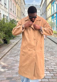 Camelfärgad dubbelknäppt kappa med stora slag, sidofickor och mjuk textur; bärs över ljusblå jeans på en kullerstensgata.
