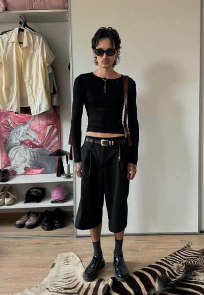 Persona delgada vestida con una camiseta de manga larga negra y corta, pantalones cortos negros, gafas de sol y zapatos de cuero, de pie sobre una alfombra de estampado de cebra en una habitación minimalista.