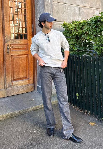 Sudadera gris y jeans negros desgastados; zapatos negros. De pie contra una puerta de madera y vegetación, con gafas en la mano.