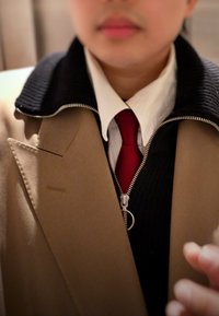 Personne portant un manteau beige par-dessus un pull à fermeture éclair noir, une chemise blanche à col et une cravate rouge, avec une main floue au premier plan.