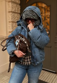 Denimová prošívaná bunda s umělým kožešinovým lemováním, tmavě hnědá semišová kabelka s kovovými doplňky a k tomu kostkovaná košile, doplněná modrými džínami.