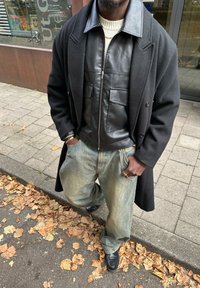 Čierna kožená bunda s prednými vreckami na krémovom svetri, vrstvená pod dlhým čiernym kabátom. Voľné svetlé džínsy a čierne topánky.