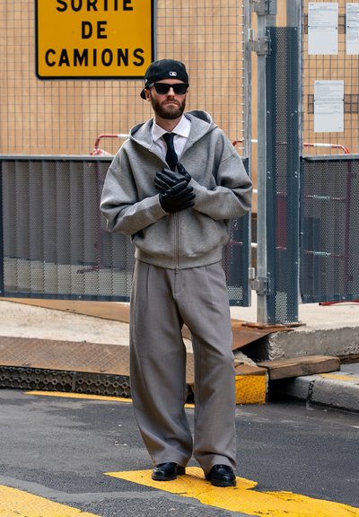 Hombre con sudadera gris con capucha, gorra negra, gafas de sol, guantes negros, corbata y pantalones holgados, de pie en la calle cerca de una valla metálica y un cartel amarillo.
