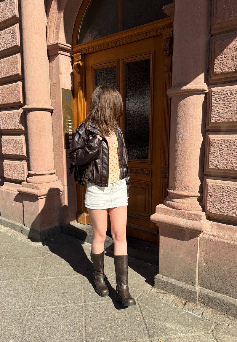 Γυναίκα που φορά καφέ δερμάτινο μπουφάν, κρεμ φούστα και μπότες στέκεται έξω από μια μεγάλη ξύλινη πόρτα σε μια ηλιόλουστη μέρα.