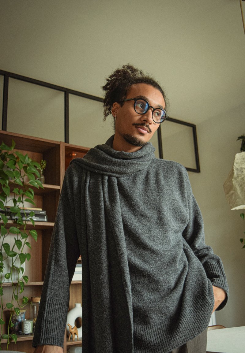 Grijze gebreide trui met een gedrapeerde halslijn, geribde mouwen en schouderdetails. Warme textuur, oversized pasvorm en casual ontwerp.