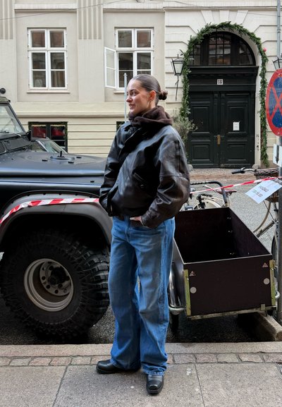 Persona con chaqueta de cuero y pantalones vaqueros anchos está en la calle junto a un vehículo todoterreno negro y una bicicleta de carga frente a una puerta arqueada.