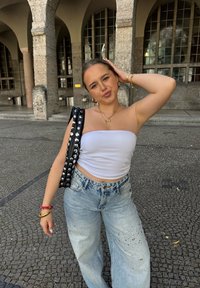 Borsa a spalla nera con borchie, top bianco senza maniche, jeans chiari strappati con decorazioni, in piedi su una superficie di ciottoli, in un contesto urbano.