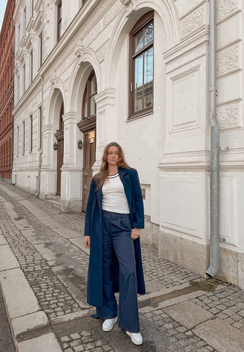 Donna giovane con cappotto lungo blu scuro e pantaloni a righe sottili in piedi su una strada di ciottoli davanti a un edificio bianco con ingressi ad arco.