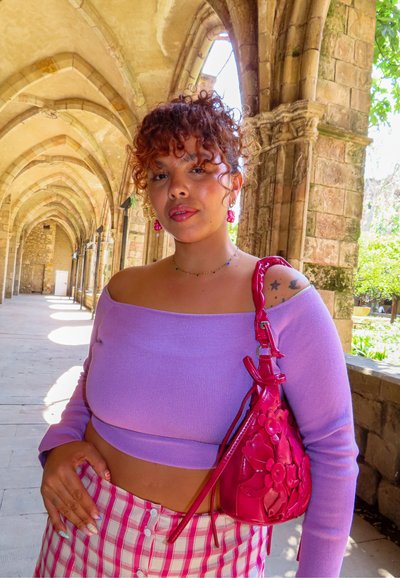 Joven mujer con cabello rizado, vistiendo un top lila de hombros descubiertos y falda de cuadros, sosteniendo un bolso floral rosa en un pasillo con arco de piedra.