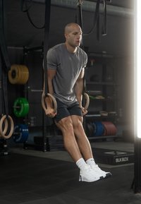 Ένα άτομο εκτελεί muscle-ups σε ξύλινες μπανέλες, φορώντας ένα γκρι μπλουζάκι και μαύρες βερμούδες, με άσπρα αθλητικά παπούτσια και άσπρες κάλτσες.