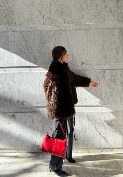 Persona con abrigo de piel oscuro y pantalones negros sosteniendo un bolso rojo, de pie frente a una pared de mármol iluminada por el sol con sombras.