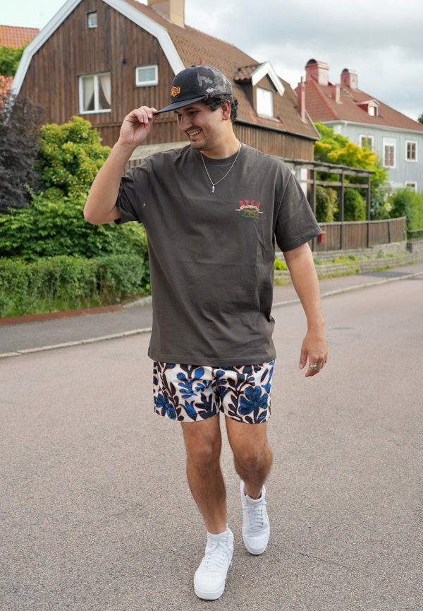 Mężczyzna uśmiecha się i idzie ulicą, ubrany w ciemny t-shirt, kwieciste szorty, białe trampki i czarną czapkę, w tle domy i zieleń.