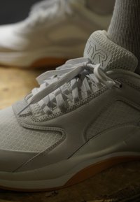 Κοντινό πλάνο λευκών sneakers με υφασμάτινη υφή και δεμένα κορδόνια, με το λογότυπο TYR στη γλώσσα, φορεμένα με ανοιχτό γκρι κάλτσες σε ξύλινο πάτωμα.