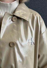 Beige, blank regnjakke med store knapper og Calvin Klein Jeans-logo. Teksturert stoff og skreddersydde kragedetaljer er synlige.