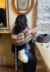 Femme en châle de fourrure et bottes blanches assise sur un tabouret près d'une fenêtre en pierre, tenant une tasse, avec une œuvre florale encadrée et un sac à pois à proximité.