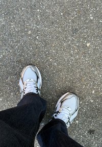 Fehér és ezüst atlétikai cipők hálós anyagból, kerek orral és fűzővel. Sötétkék farmerral viseletben, egy texturált szürke felület előtt.