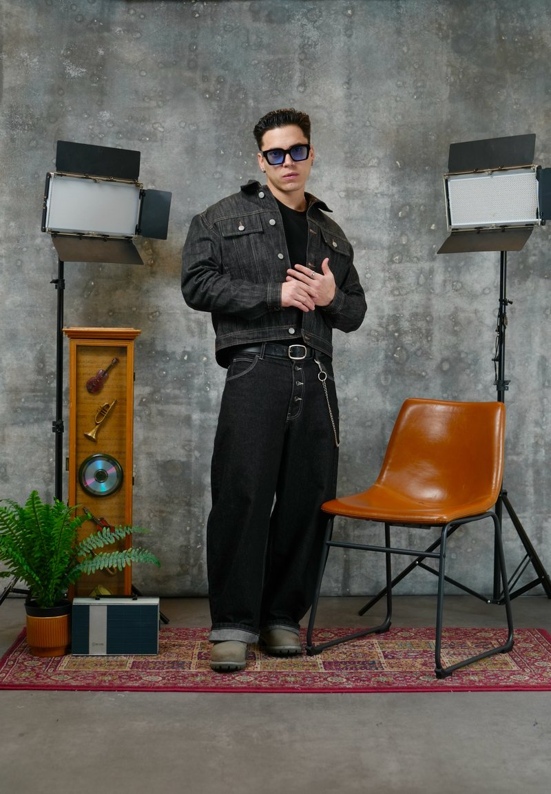 Mann in dunkler Jeansjacke und weitem Jeans steht auf einem gemusterten Teppich zwischen Studiolampen, einem braunen Stuhl, einer Topfpflanze und einem Vintage-Lautsprecher.