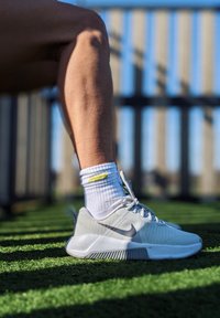 Šviesiai pilki sportiniai batai su tekstūruota viršutine dalimi, su baltais raišteliais ir juodu Nike ženklu, dėvimi su balta kojinėmis su geltonu akcentu.