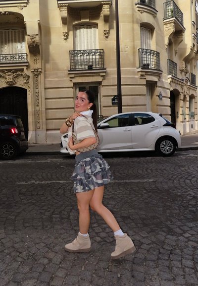 Figura femenina vestida con una blusa de rayas, una falda con patrón de camuflaje y botas de tobillo beige, llevando un bolso estampado frente a un fondo urbano.