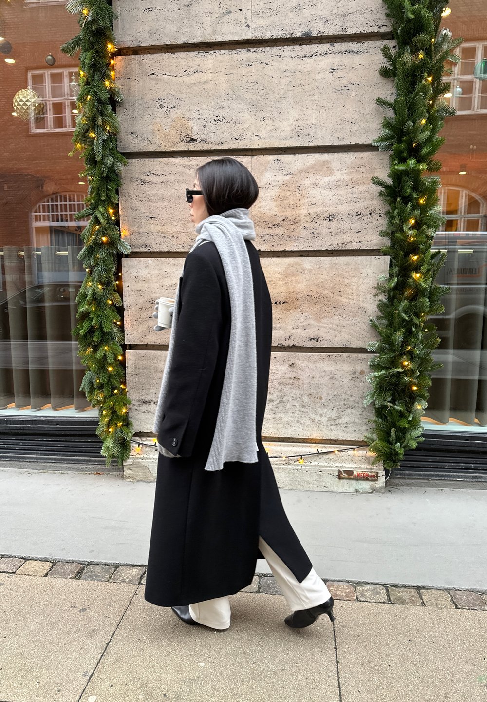 Sort lang frakke, gråt tørklæde, cremefarvede bukser og sorte sko; stående ved en struktureret stenvæg med grønt og lys. Kaffekop i hånden.