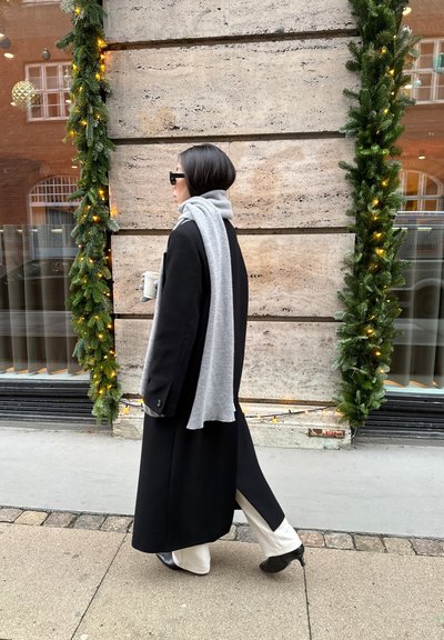 Abrigo largo negro, bufanda gris, pantalones crema y zapatos negros; de pie junto a una pared de piedra texturizada con vegetación y luces. Taza de café en mano.