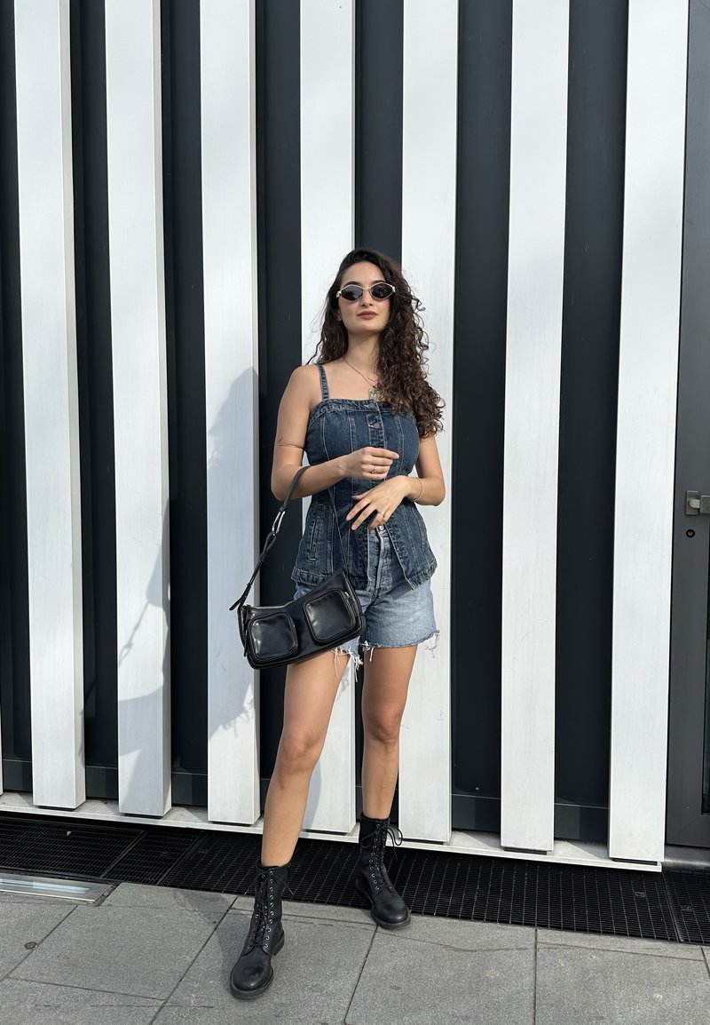 Donna con top e shorts in denim, stivali neri e borsa, in piedi davanti a un muro a righe verticali bianche e nere.