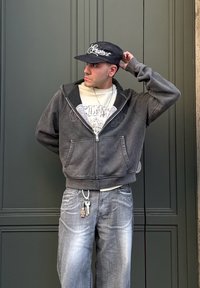 Felpa con cappuccio grigio scuro con cerniera, sotto una t-shirt grafica grigio chiaro. Abbinata a jeans grigio sbiadito e dettagli con portachiavi. Cappellino nero con scritte bianche.