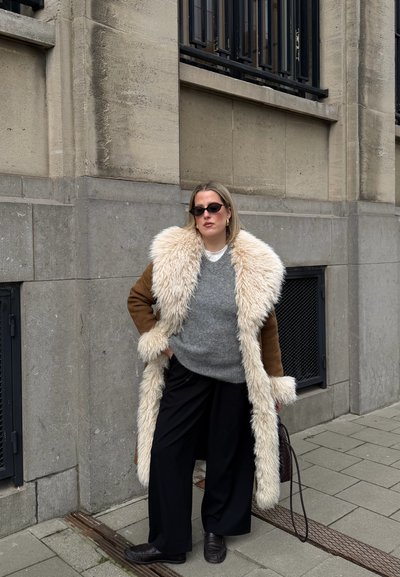 Mujer con un abrigo largo marrón con un gran ribete de piel crema, suéter gris, pantalones negros y gafas de sol, de pie en la acera de la ciudad contra una pared de piedra.