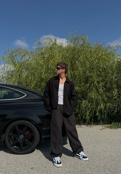 Chaqueta negra, camiseta blanca sin mangas y pantalones sueltos negros. Zapatillas negras con cordones blancos. De pie al lado de un coche negro y con vegetación en el fondo.