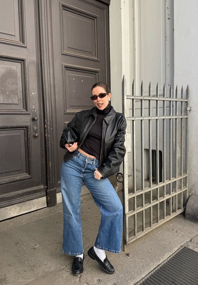 Mujer con chaqueta de cuero negra, jersey de cuello vuelto negro, vaqueros de pernera ancha, mocasines negros y pequeño bolso negro, posando junto a una gran puerta de madera y una reja de metal.