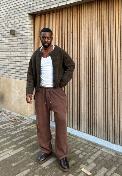 Cardigan de canalé marrón sobre una camiseta blanca, combinado con unos pantalones holgados marrones y zapatos oscuros sin cordones. El fondo presenta paneles de madera texturizados.